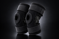 A pair of black knee support braces with adjustable straps and a quick-release button, displayed on a dark reflective studio background.